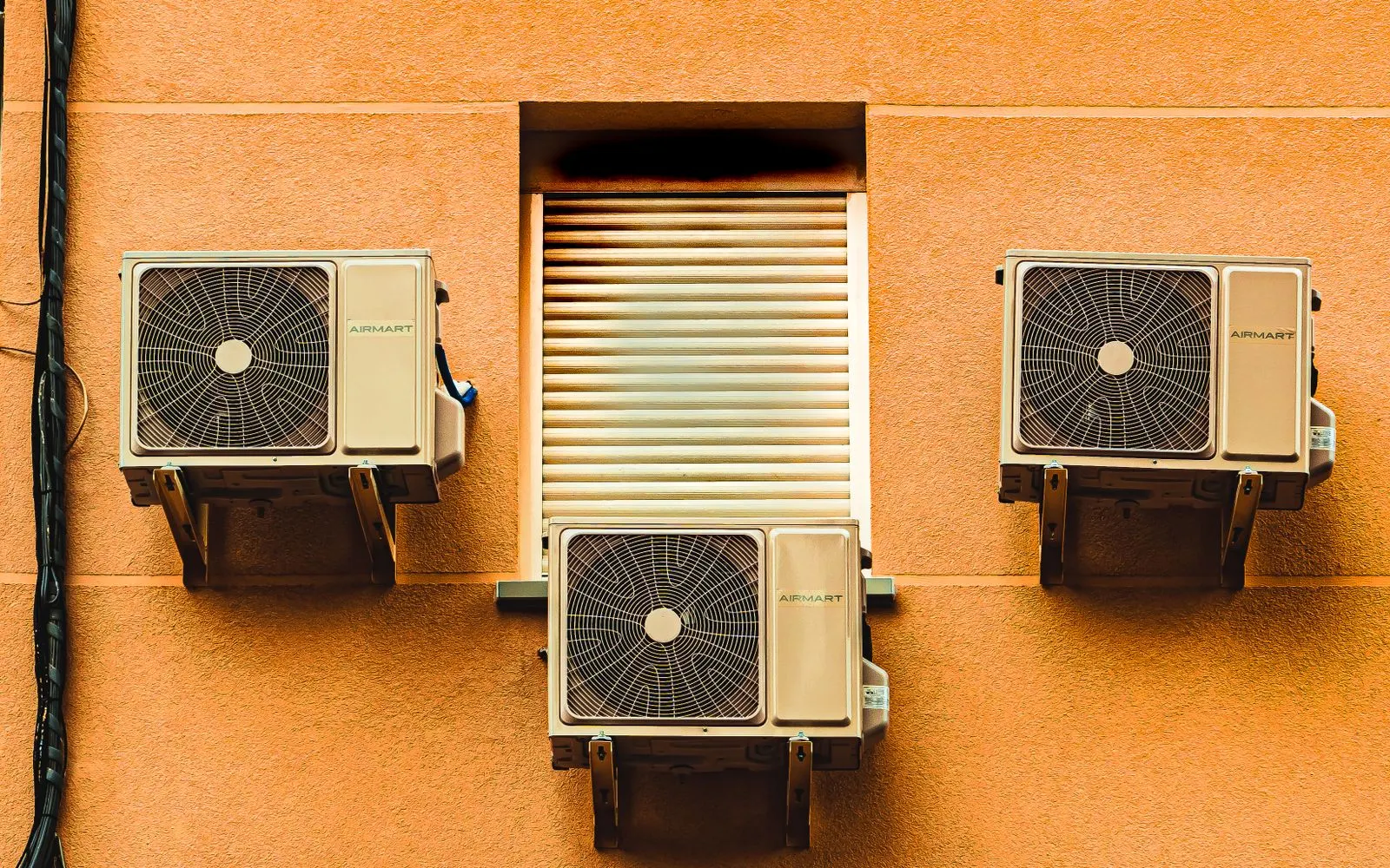 Trois unités extérieures de climatiseur fixées sur la façade orangée d'un immeuble, de part et d'autre d'un volet roulant fermé