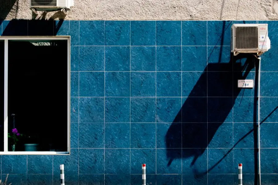 Unité extérieure de climatiseur fixée sur un mur carrelé bleu, avec son ombre projetée au soleil